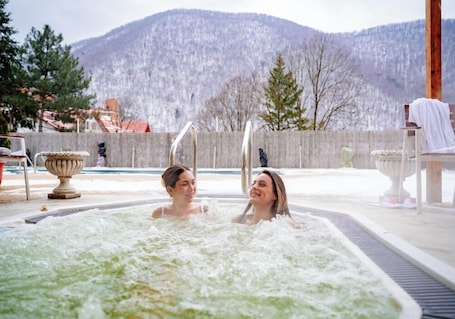 A hot tub with two people, a stone wall, and a mountain in the background.