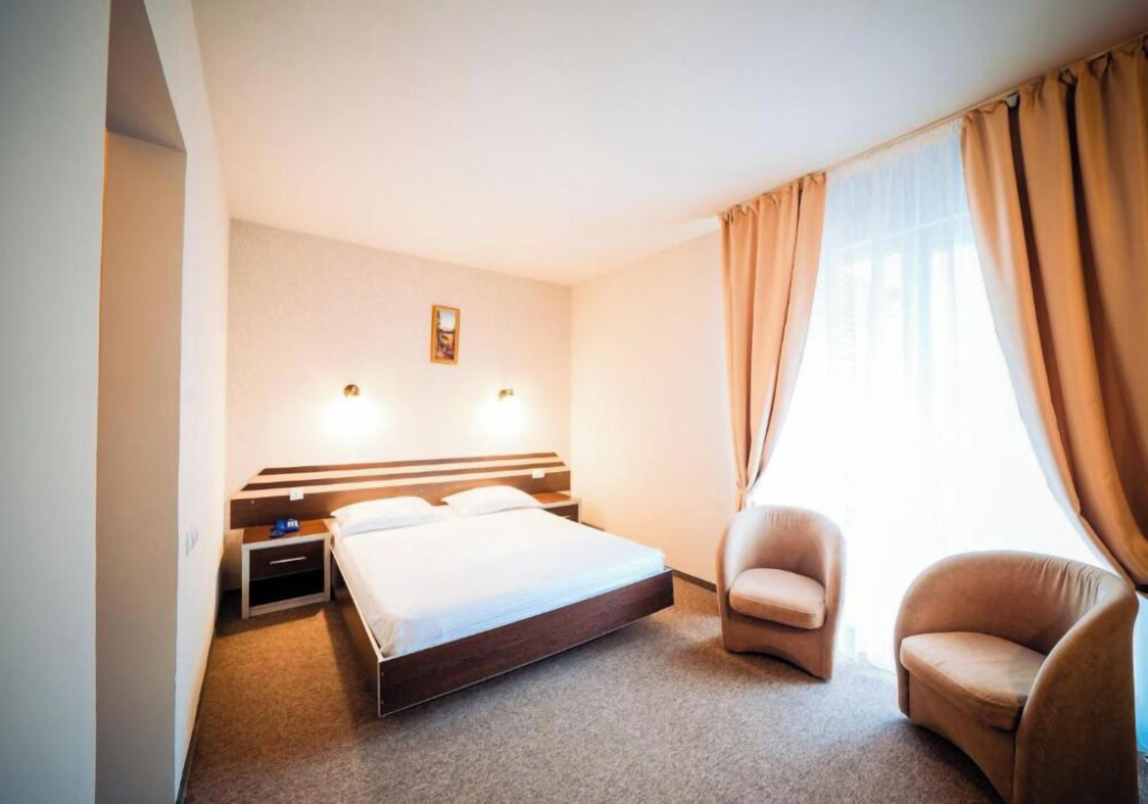 A hotel room with a bed, two chairs, a nightstand, and a wall-mounted light.