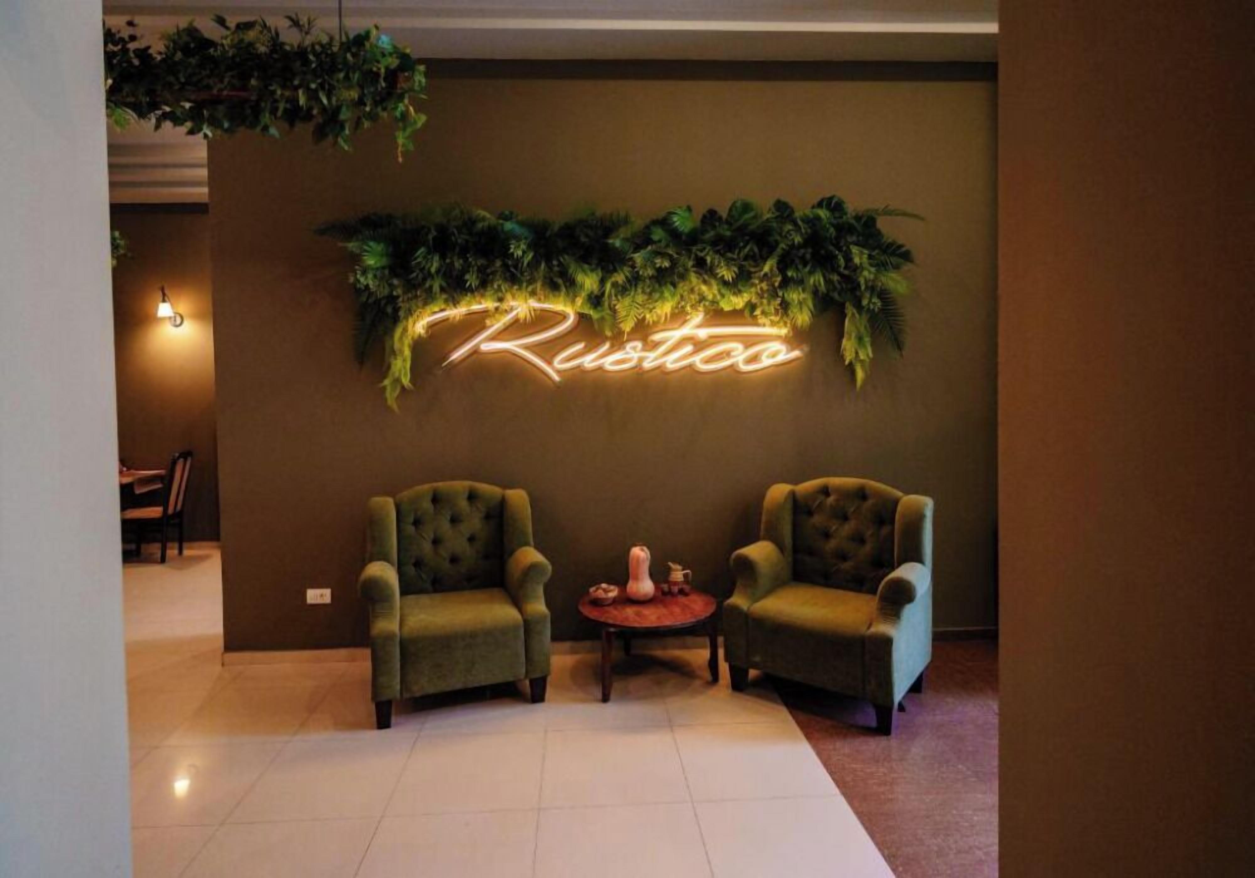 Eine Lobby mit zwei grünen Sesseln, einem kleinen runden Tisch, einem leuchtenden 'Rustico'-Schild und einer Wandpflanze.