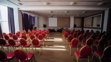 Un salón de conferencias con hileras de sillas rojas, una pantalla de proyector y un espejo grande en la pared.