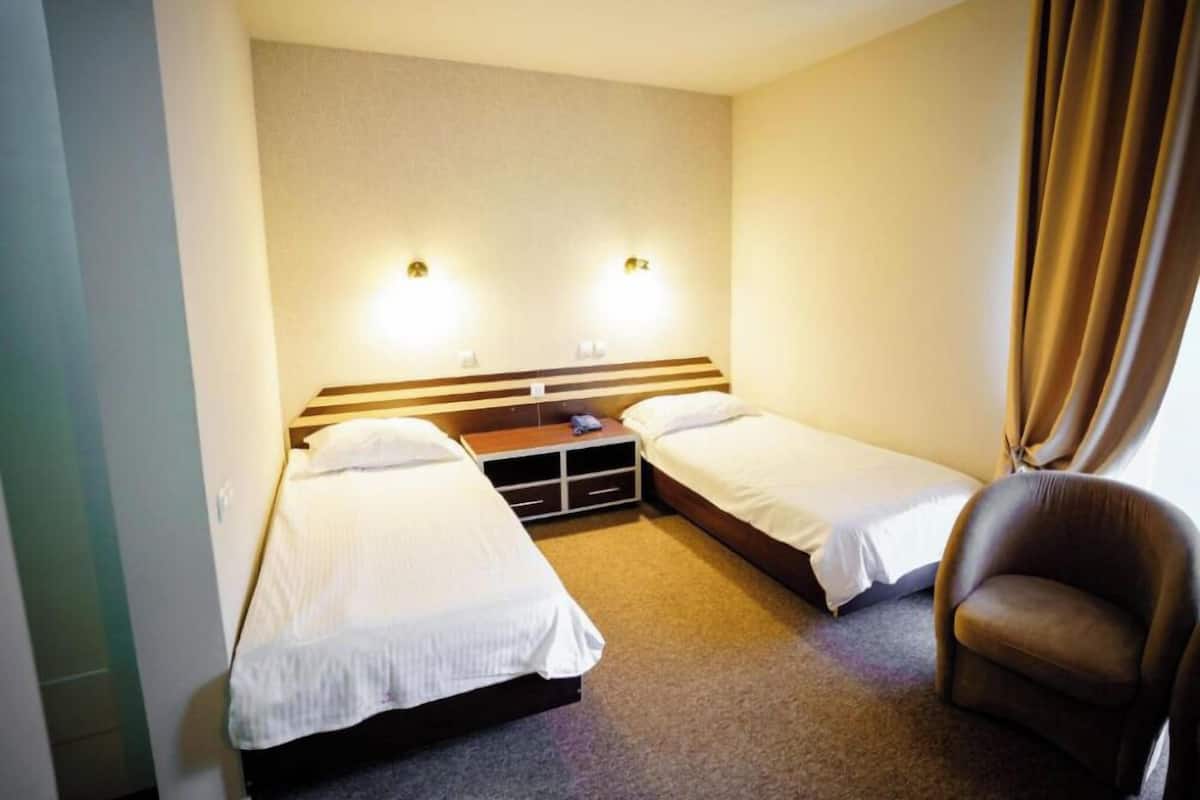 A hotel room with two single beds, a wooden headboard, a small table, a chair, and two wall-mounted lights.