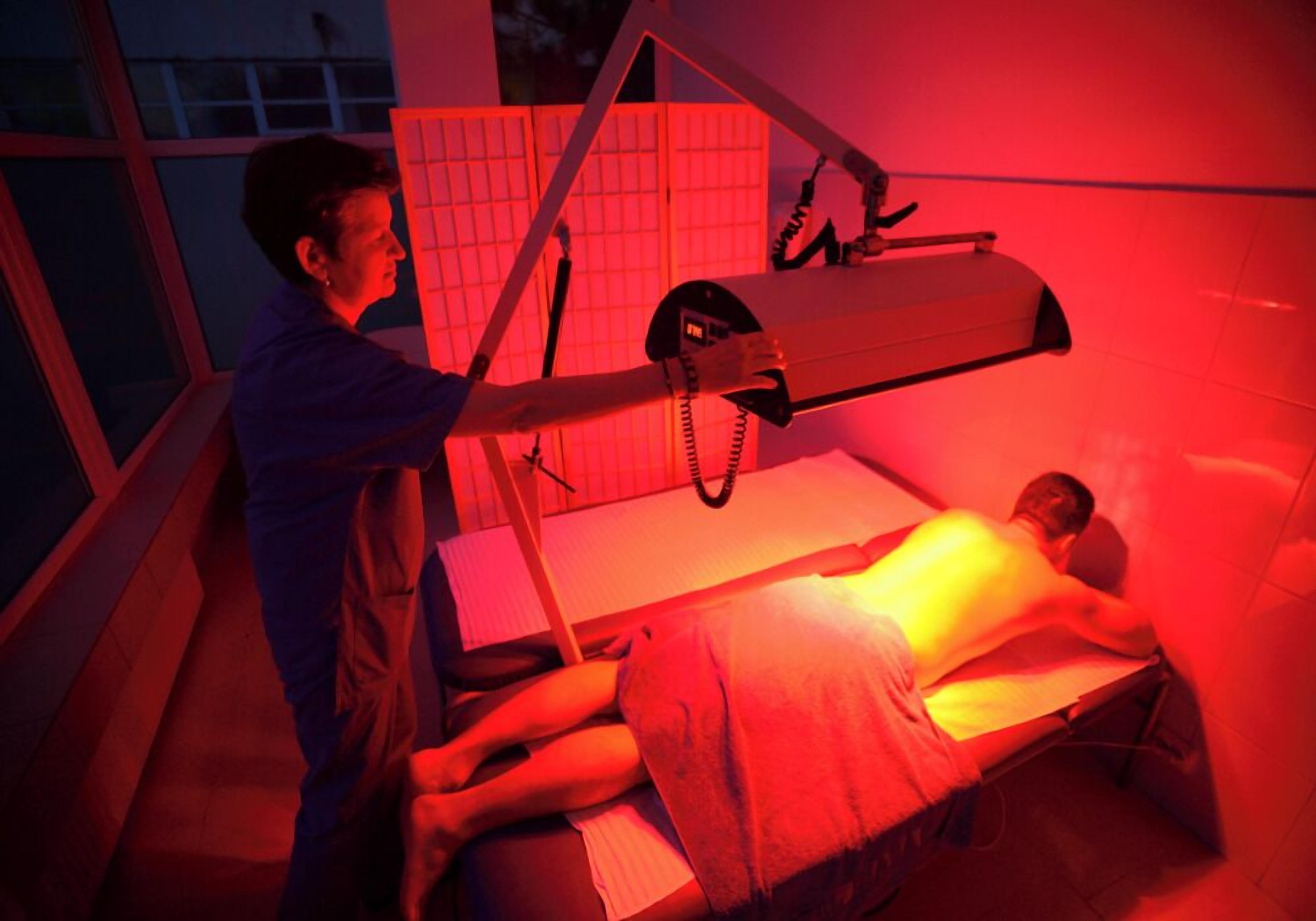 A medical procedure room with a patient lying on a bed, a medical professional standing beside, and a large overhead lamp.