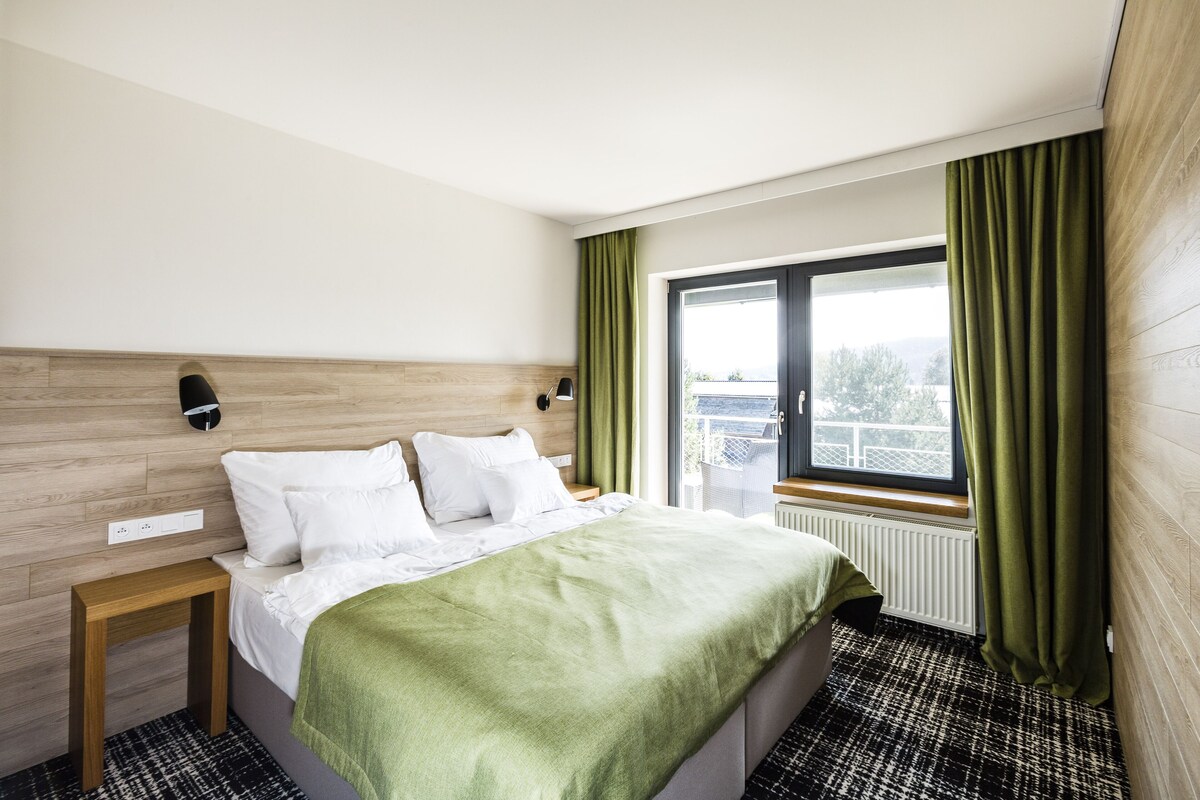 A hotel room with a bed, a nightstand, a window with curtains, and a view of the outdoors.
