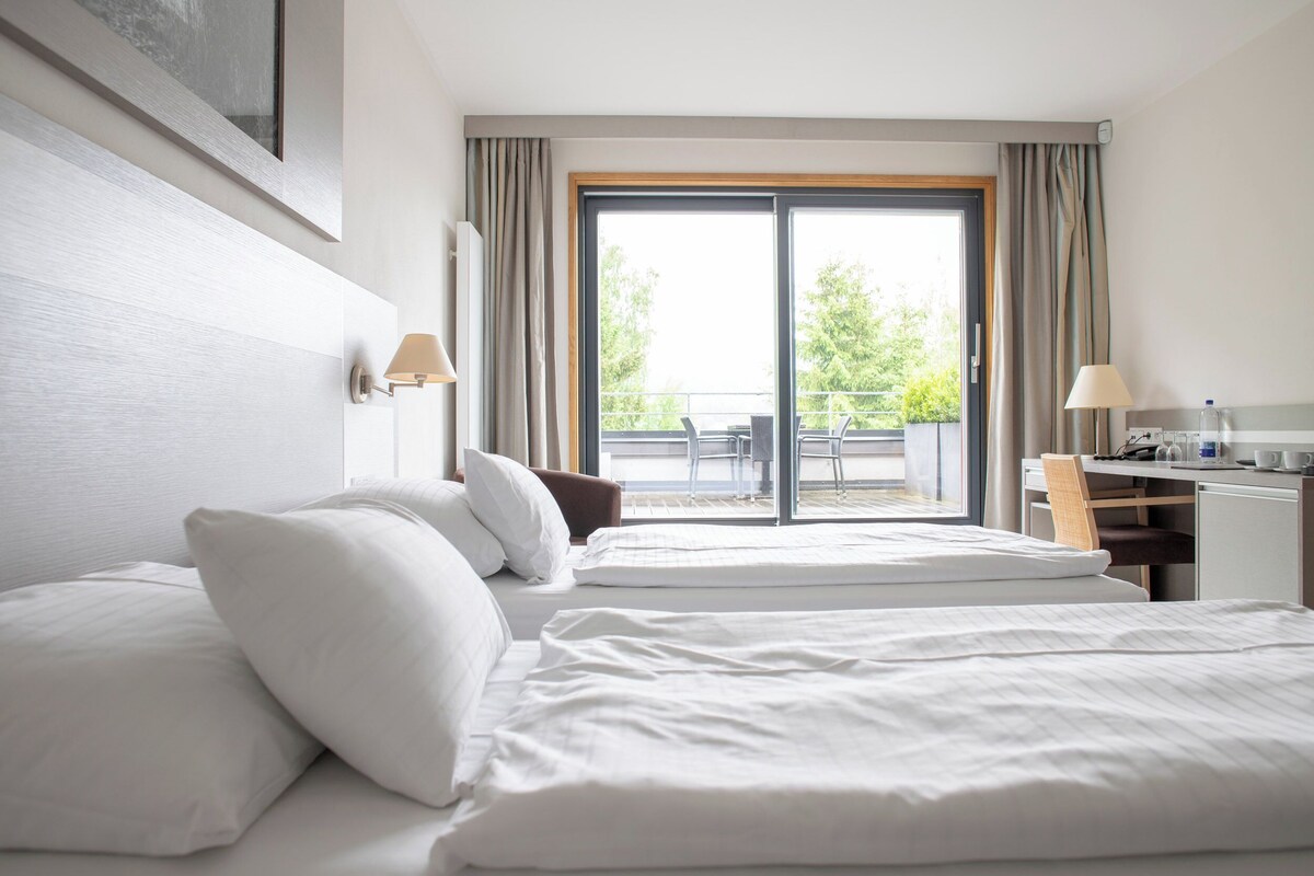 A hotel room with two beds, a desk, a chair, and a balcony with a view of greenery.