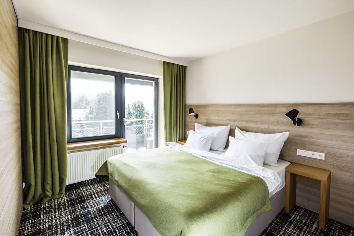 A hotel room with a bed, a nightstand, a window with curtains, and a view of trees outside.