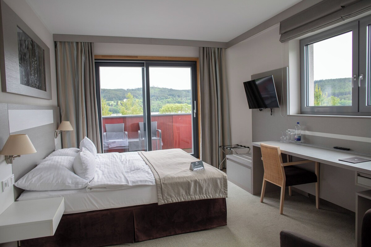 A hotel room with a bed, desk, chair, TV, and a balcony with a view of greenery.
