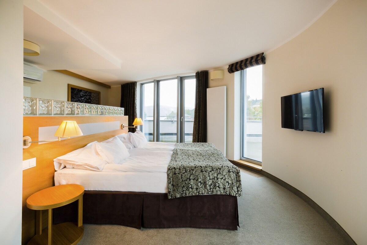 A hotel room with a bed, bedside table, lamp, TV, and large windows.