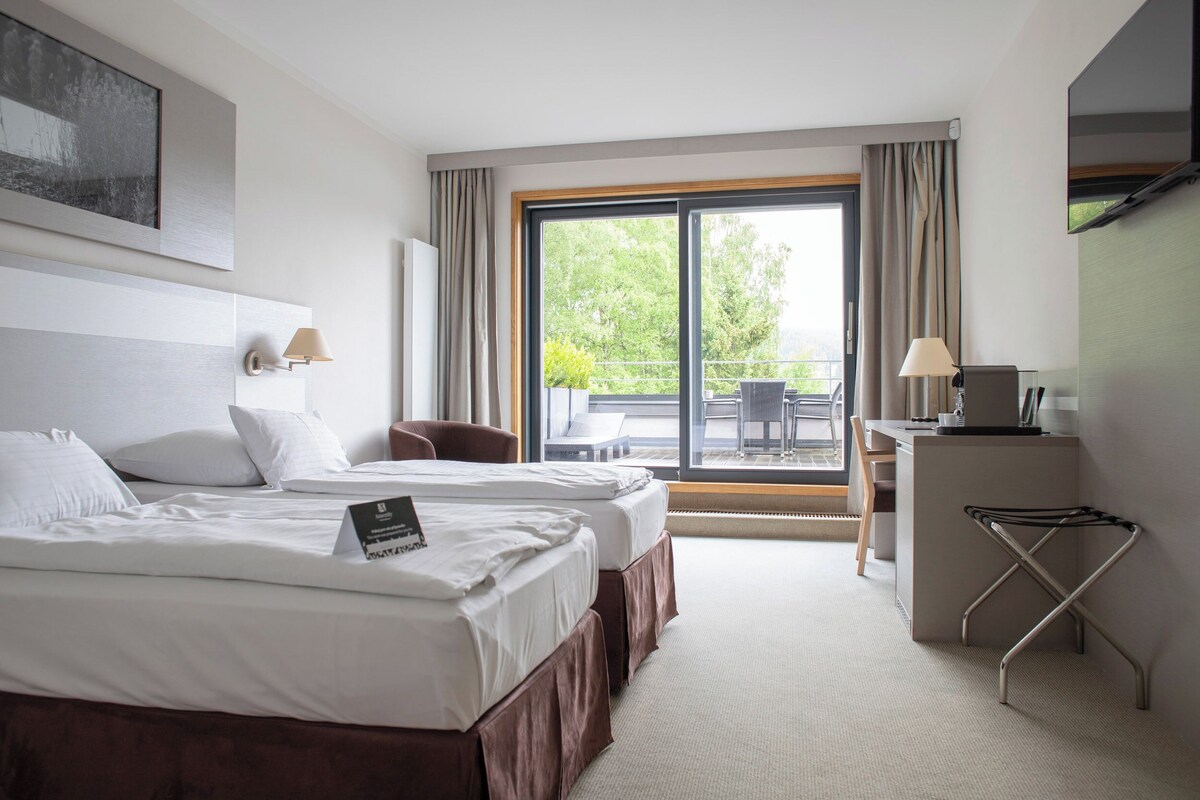 A hotel room with two beds, a desk, a chair, and a balcony with a view of greenery.