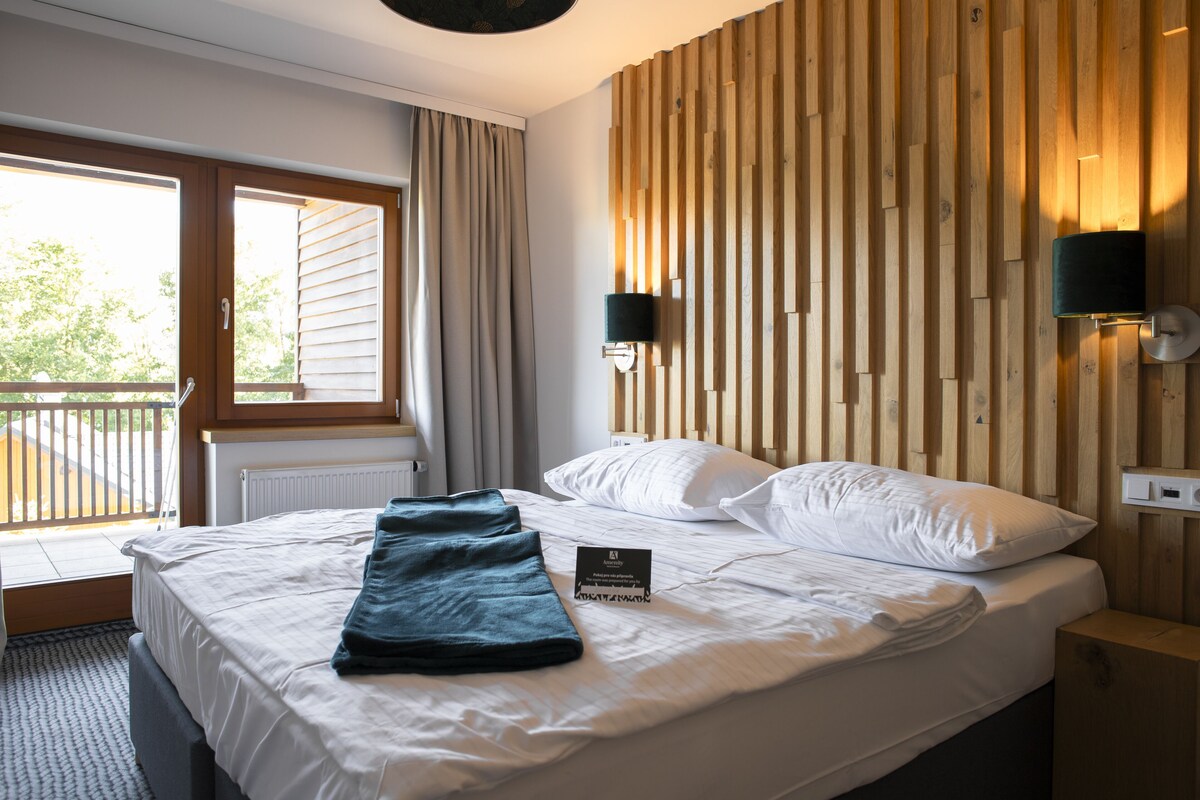 A hotel room with a bed, a window with curtains, a wooden wall panel, and a wall-mounted light fixture.