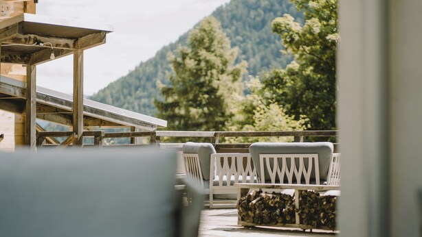 A wooden deck with white railings, a seating area with cushions, and a view of a forested mountain. - Warm vacation apartment at Hof Neuhaus (Platzers)