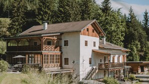 A traditional chalet with a wooden balcony, a stone staircase, and a view of a forested hillside. - Warm vacation apartment at Hof Neuhaus (Platzers)