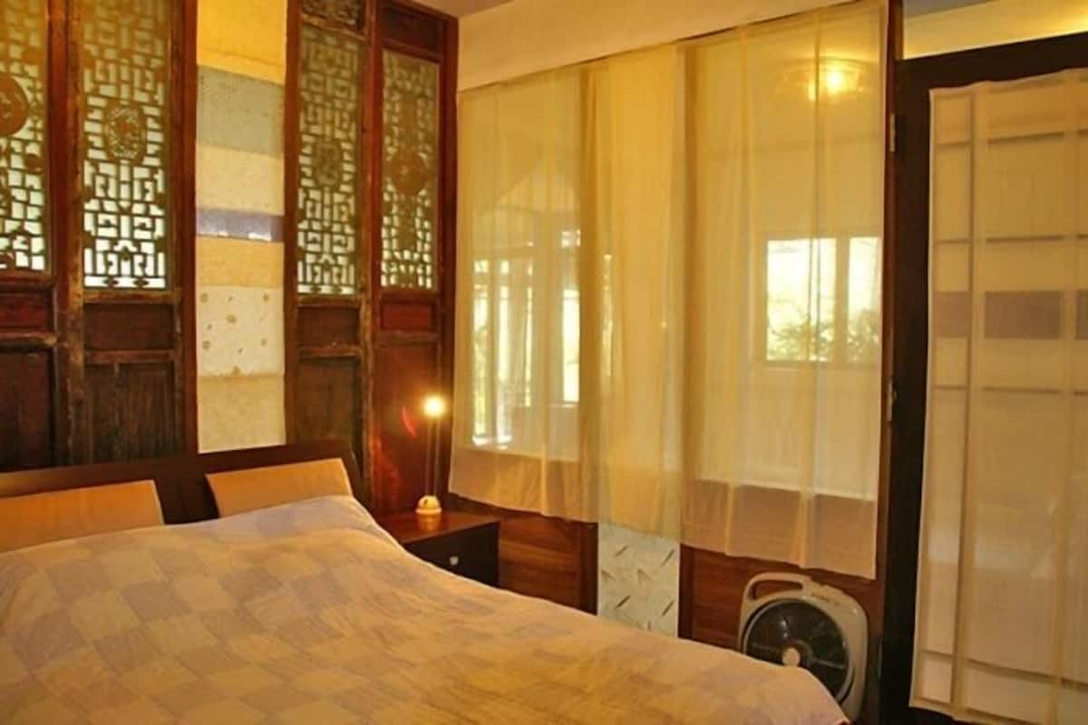 A traditional room with a bed, wooden paneling, a window with sheer curtains, and a wall-mounted fan.