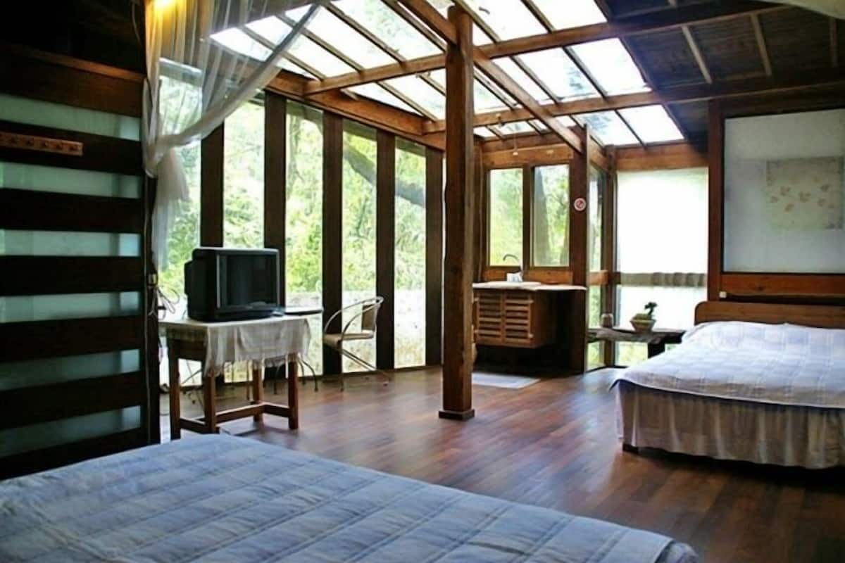 A room with two beds, a desk with a television, and large windows allowing natural light.