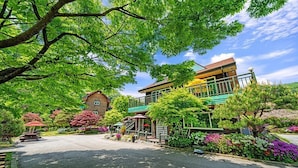 A two-story building with a green roof and a balcony, surrounded by lush greenery and flowering plants.