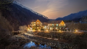 A two-story wooden lodge with a stone pathway leading to a small stream, surrounded by mountains and lit by warm lights.