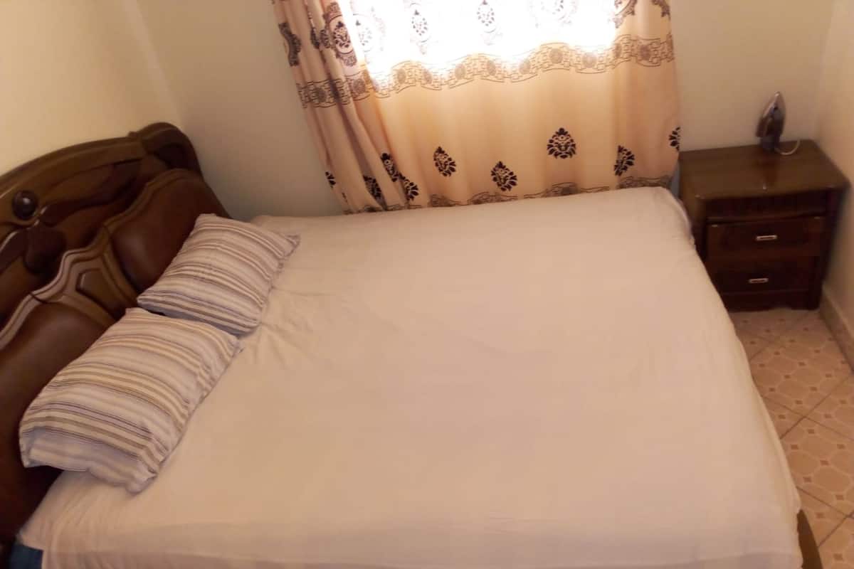 Uma cama bem arrumada com cabeceira de madeira, uma pequena mesa de cabeceira com abajur e cortina estampada.