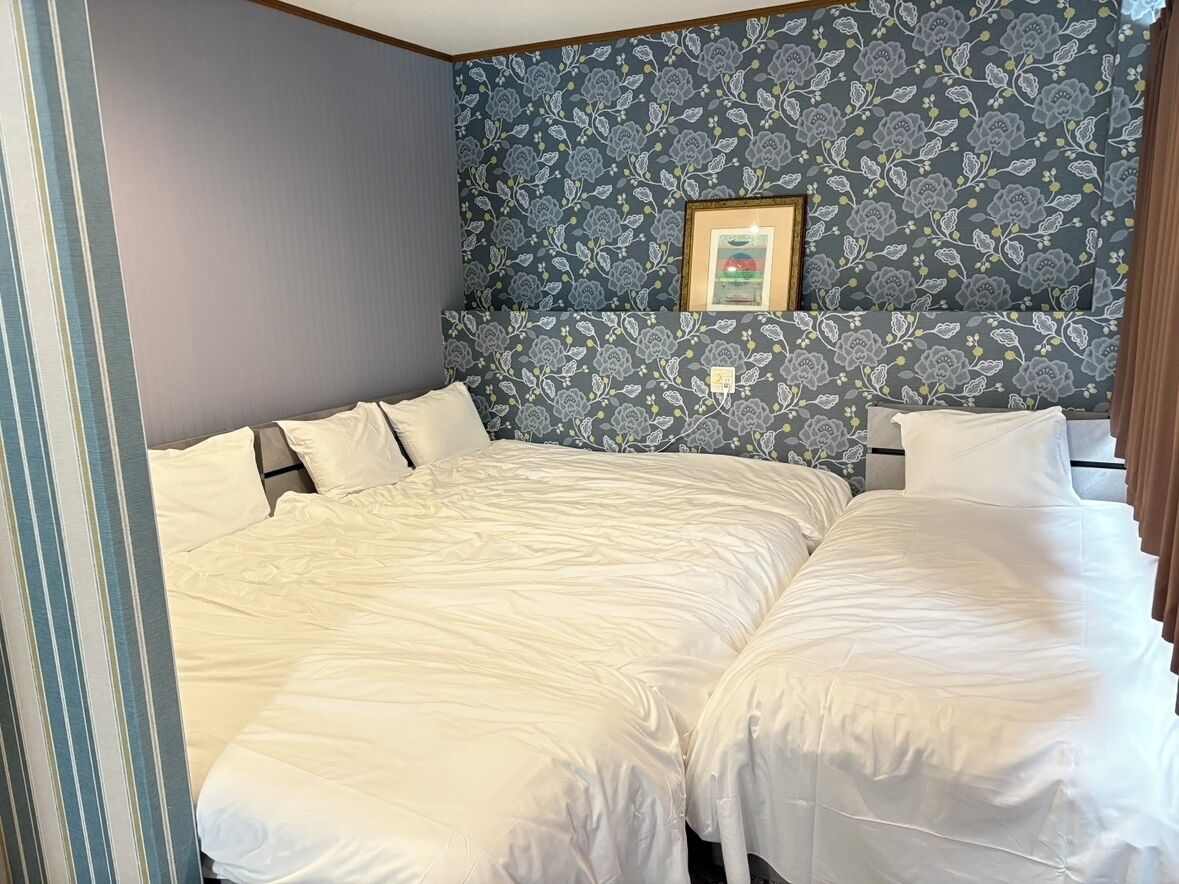 Un lit double avec une tête de lit adossée à un mur orné d’un motif floral, un tableau encadré au-dessus, et une fenêtre avec des rideaux.