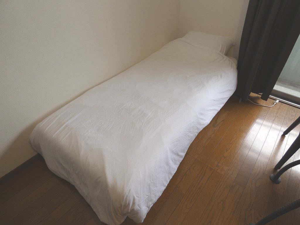 A small, single-bed room with a white bedspread, wooden flooring, and a window with curtains.