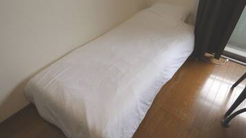 A small, single-bed room with a white bedspread, wooden flooring, and a window with curtains.