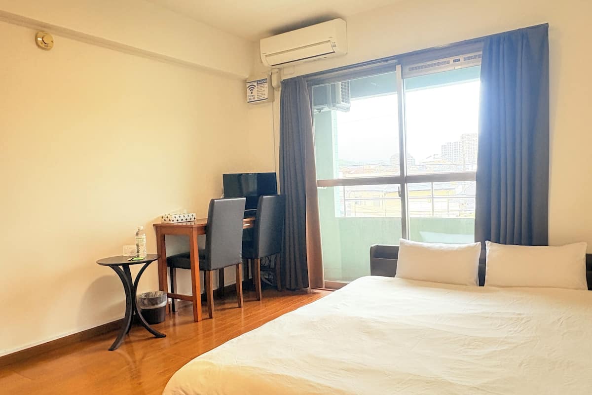 Een hotelkamer met een bed, een bureau met stoel, een tafeltje met een fles en een raam met gordijnen.