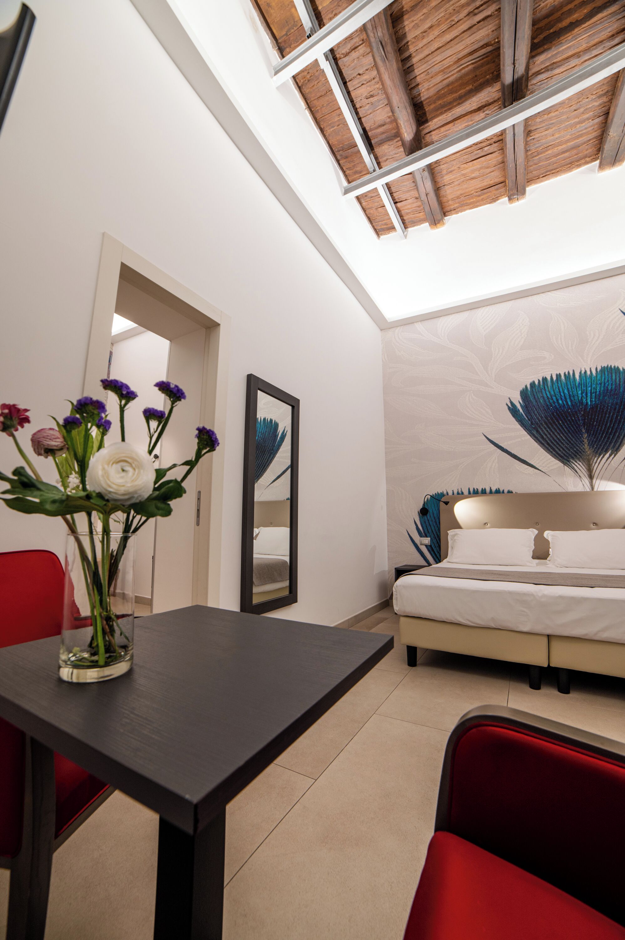 A modern bedroom with a skylight, a bed with a headboard, a bedside table, a vase with flowers, and a red chair.