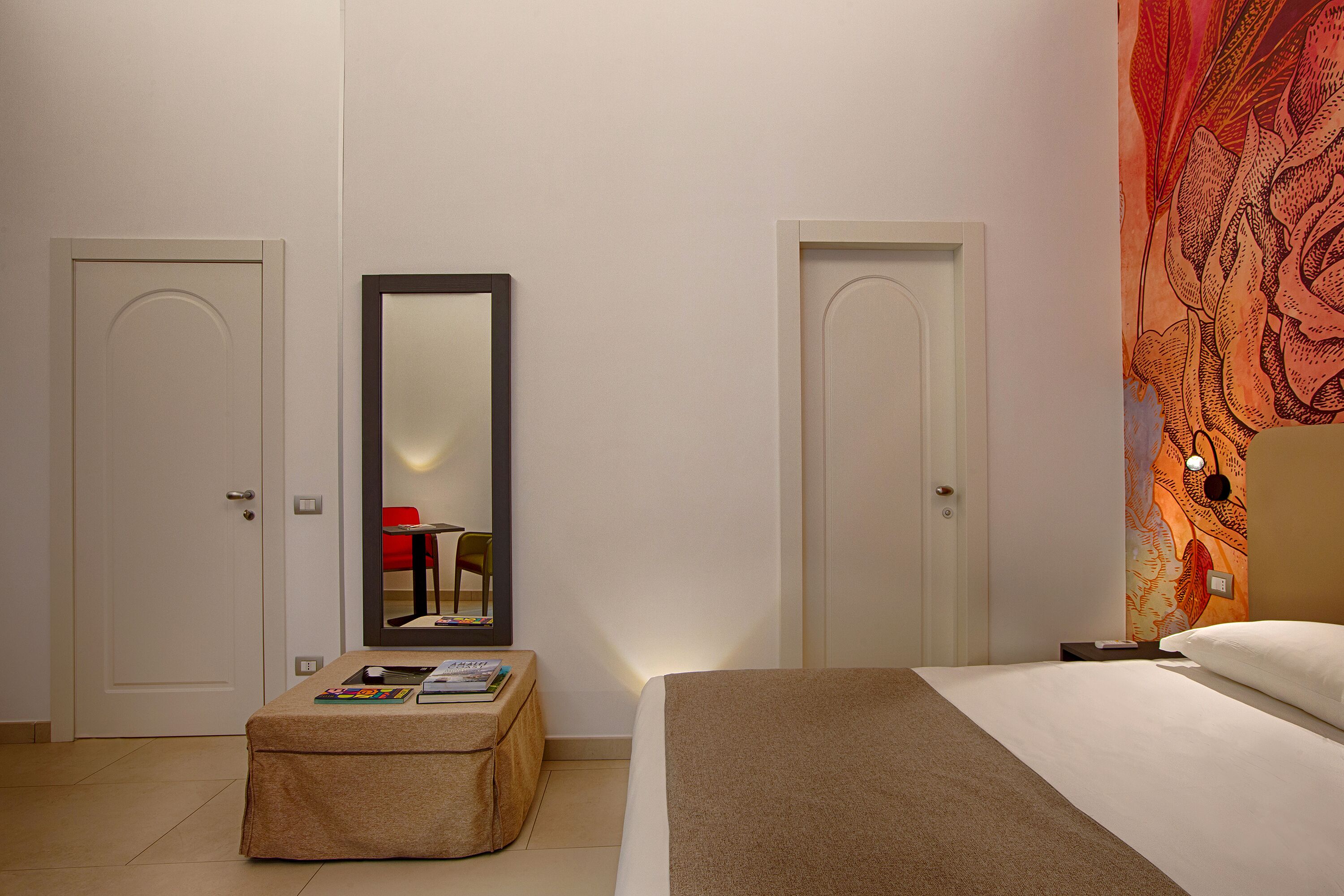 Quarto de hotel com cama, mesa de cabeceira com luminária, espelho e parede com tapeçaria floral.