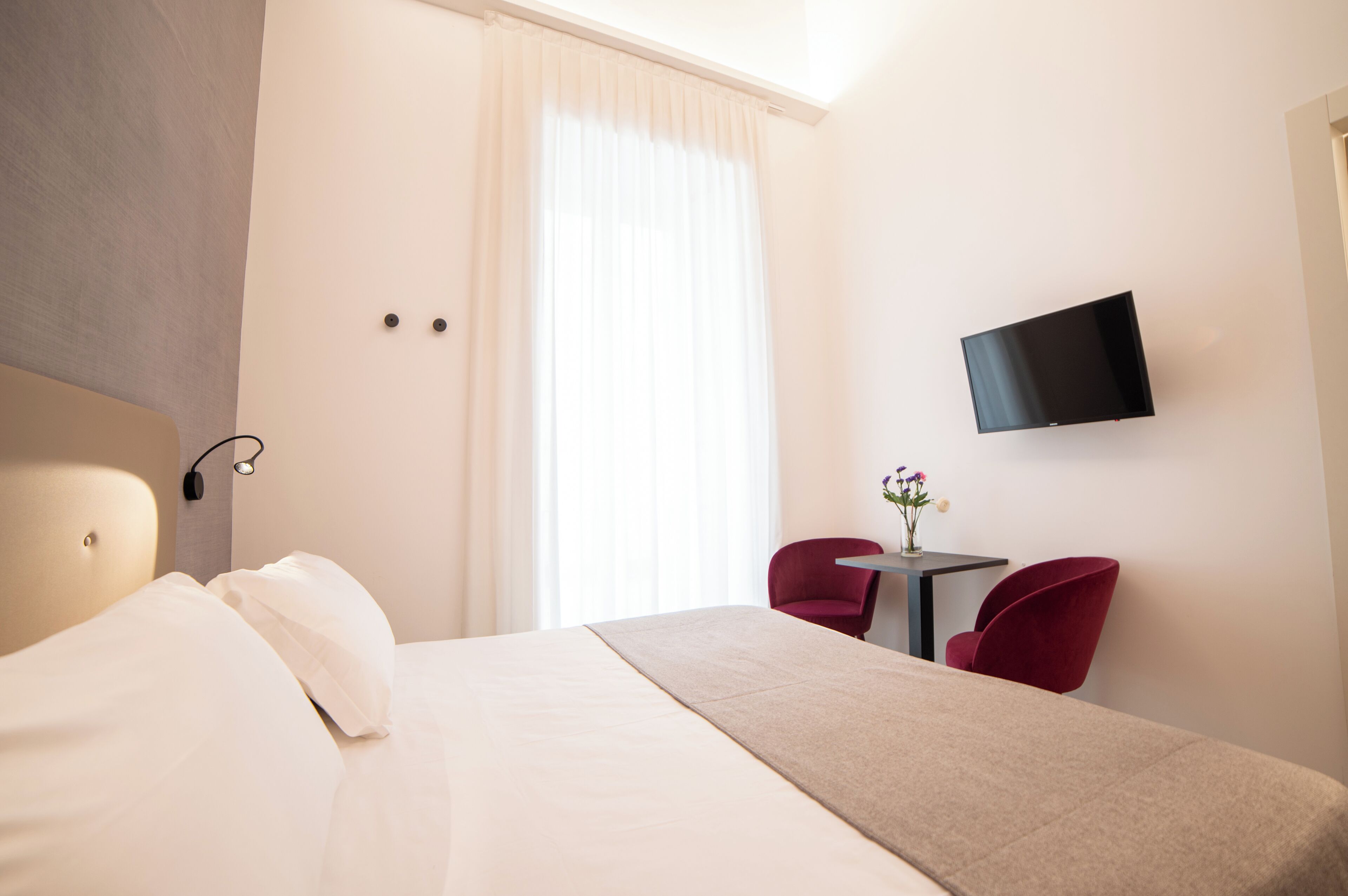 A hotel room with a bed, a TV mounted on the wall, a small table with a vase of flowers, and two red chairs.