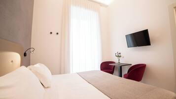 A hotel room with a bed, a TV mounted on the wall, a small table with a vase of flowers, and two red chairs.