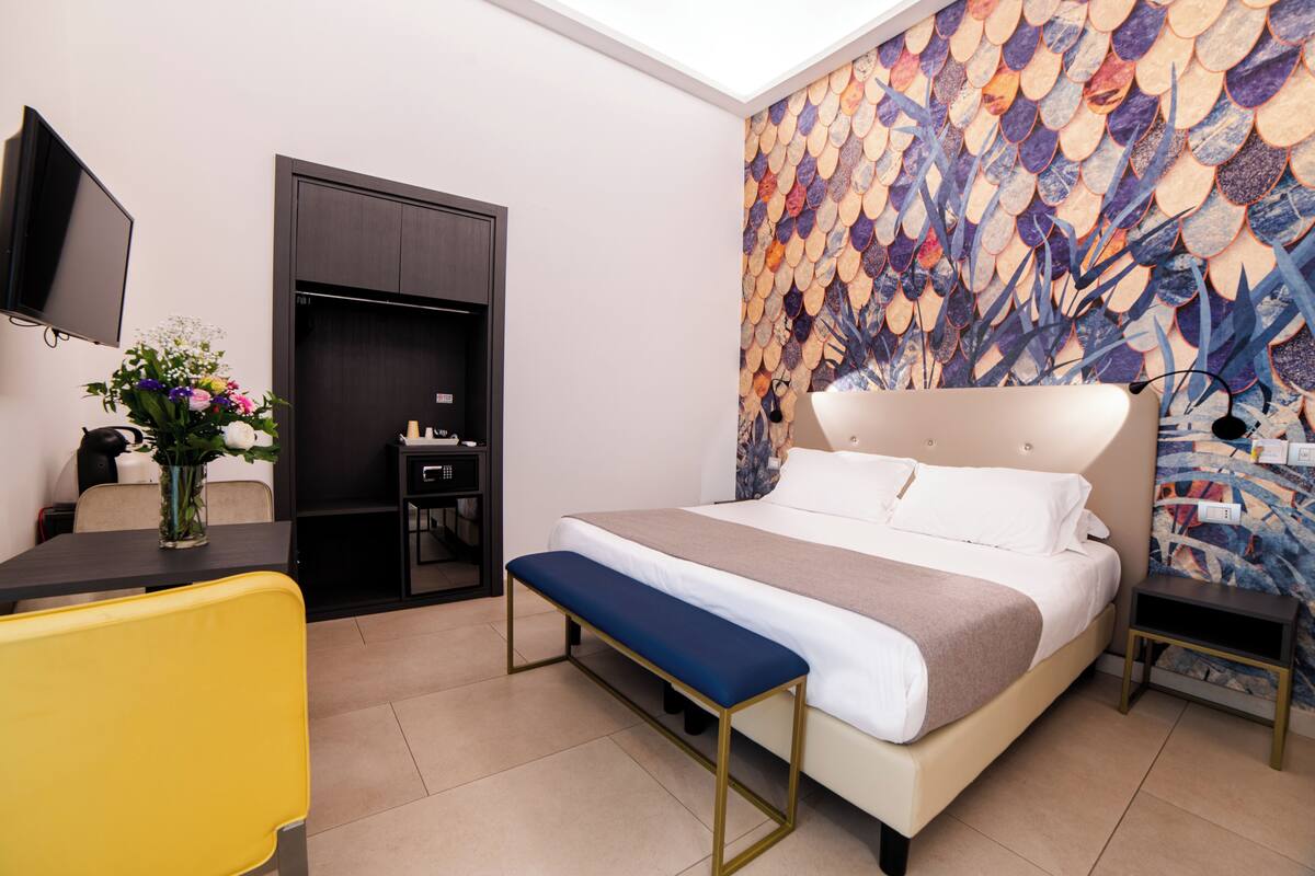 A modern hotel room with a large bed, a desk, a chair, a TV, and a decorative wall.