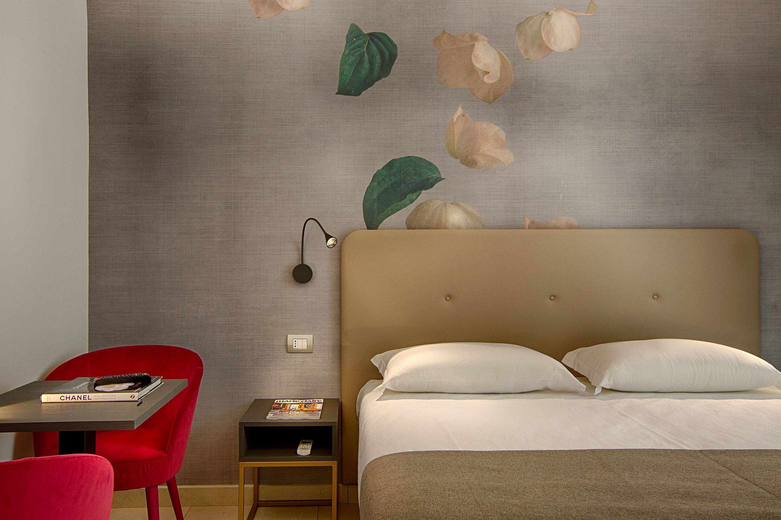 Habitación de hotel con una cama, una silla roja, una mesita pequeña y una pared con un mural floral.