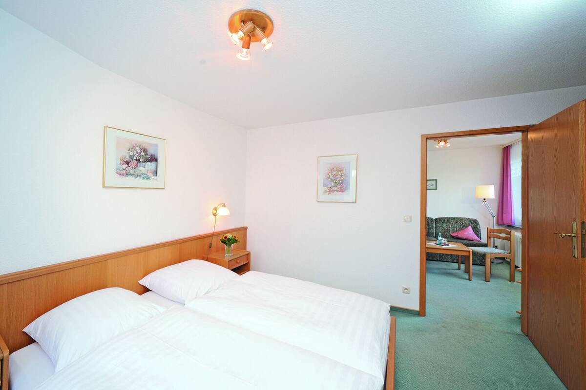 A hotel room with a double bed, a nightstand with a lamp, two framed pictures on the wall, and a view into the adjacent room through an open door.