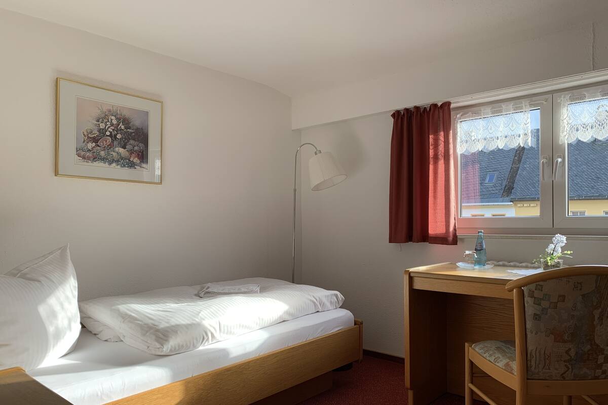 A single bed with a wooden frame, a desk with a chair, a floor lamp, and a window with curtains.