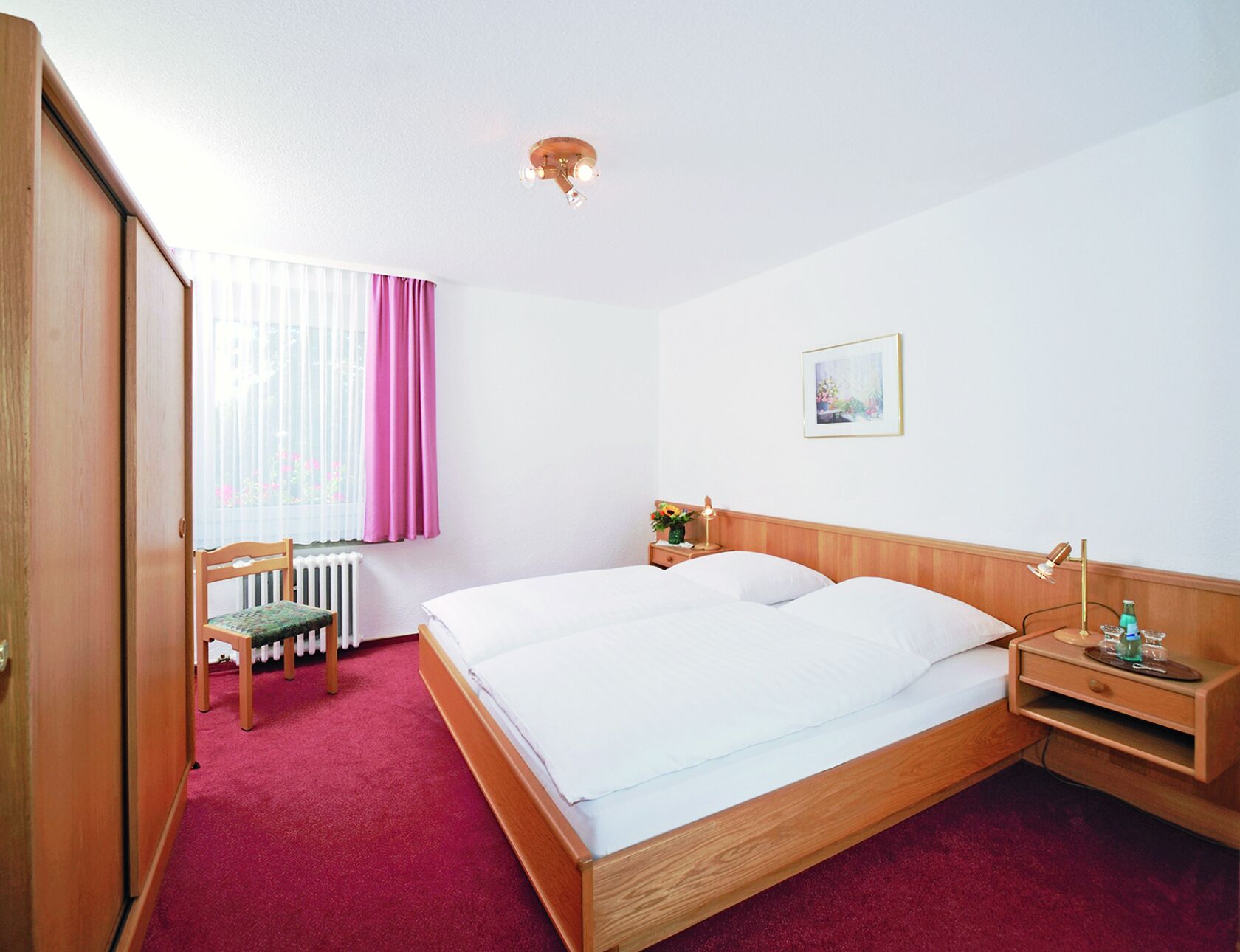 Ein Hotelzimmer mit einem hölzernen Bett, einem Sessel, einem Nachttisch und einem Fenster mit Vorhängen.