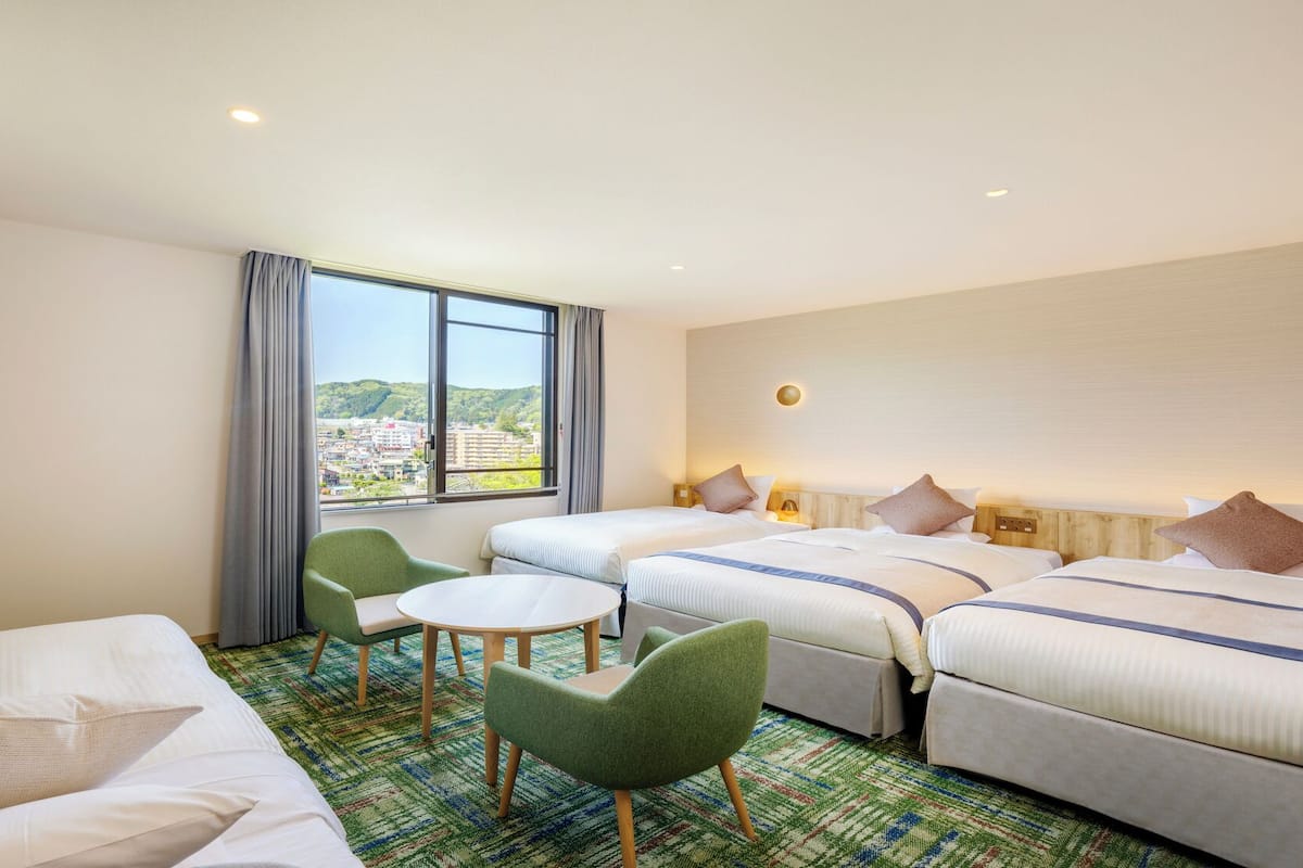 A hotel room with two beds, a small table, and chairs. There is a window with a view of buildings and greenery.
