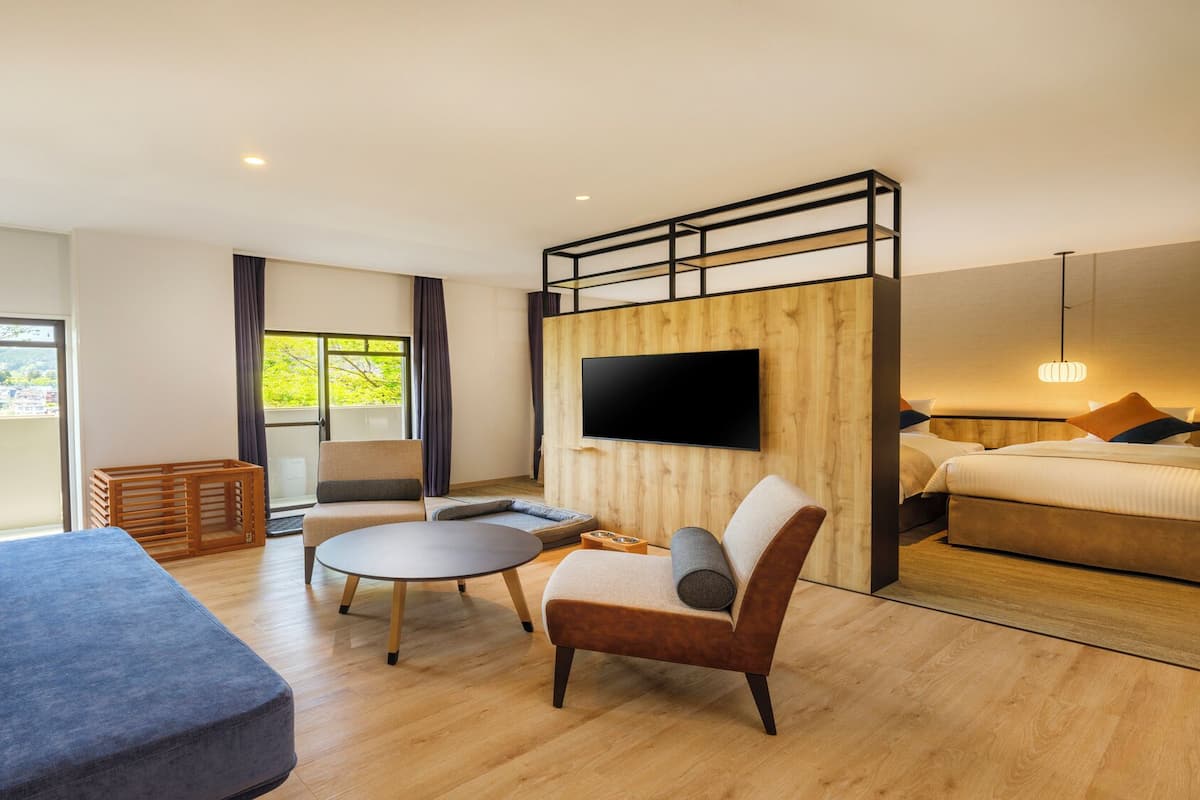 A modern hotel room with a sofa, armchair, and a small round table. There is a TV mounted on a wooden wall, and a bed with pillows.
