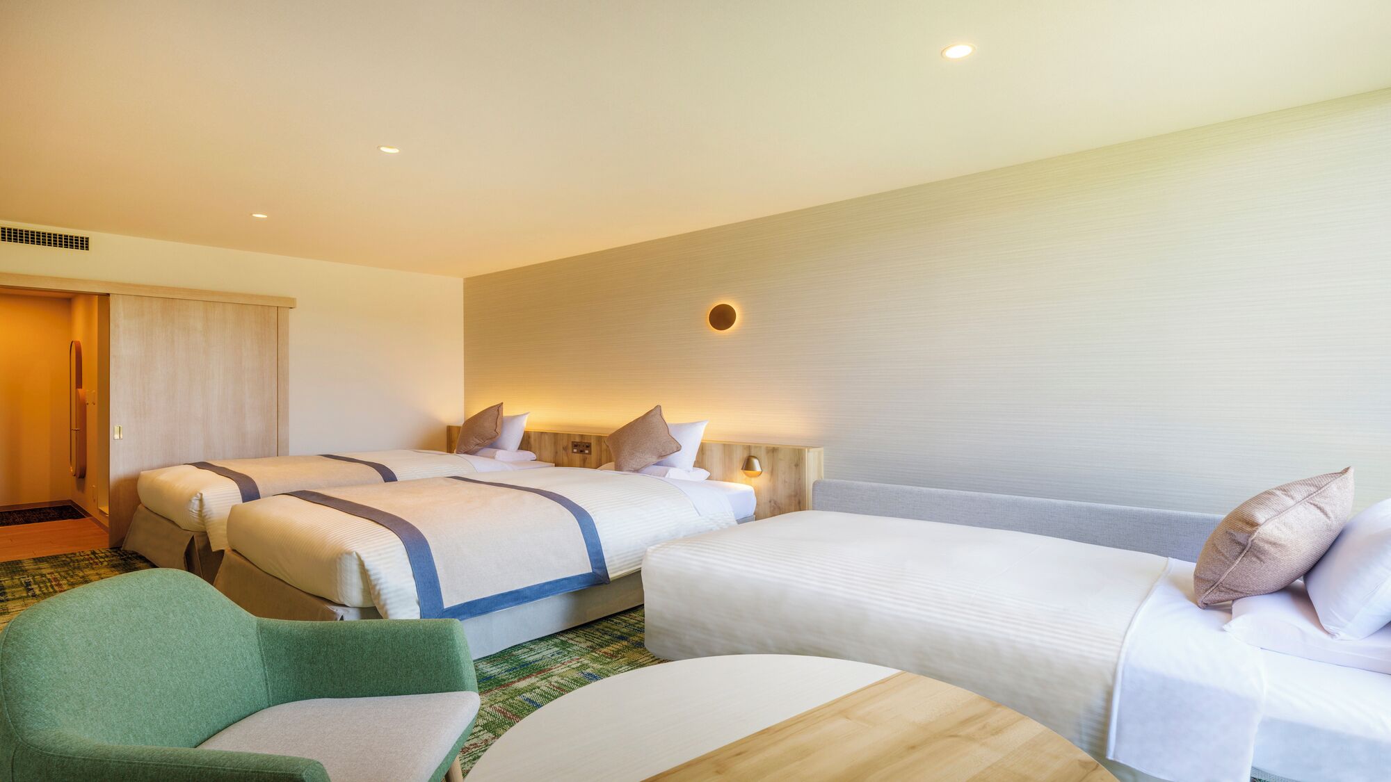 Une chambre d’hôtel avec deux lits simples, un fauteuil vert, une table d’appoint en bois et une petite table ronde.
