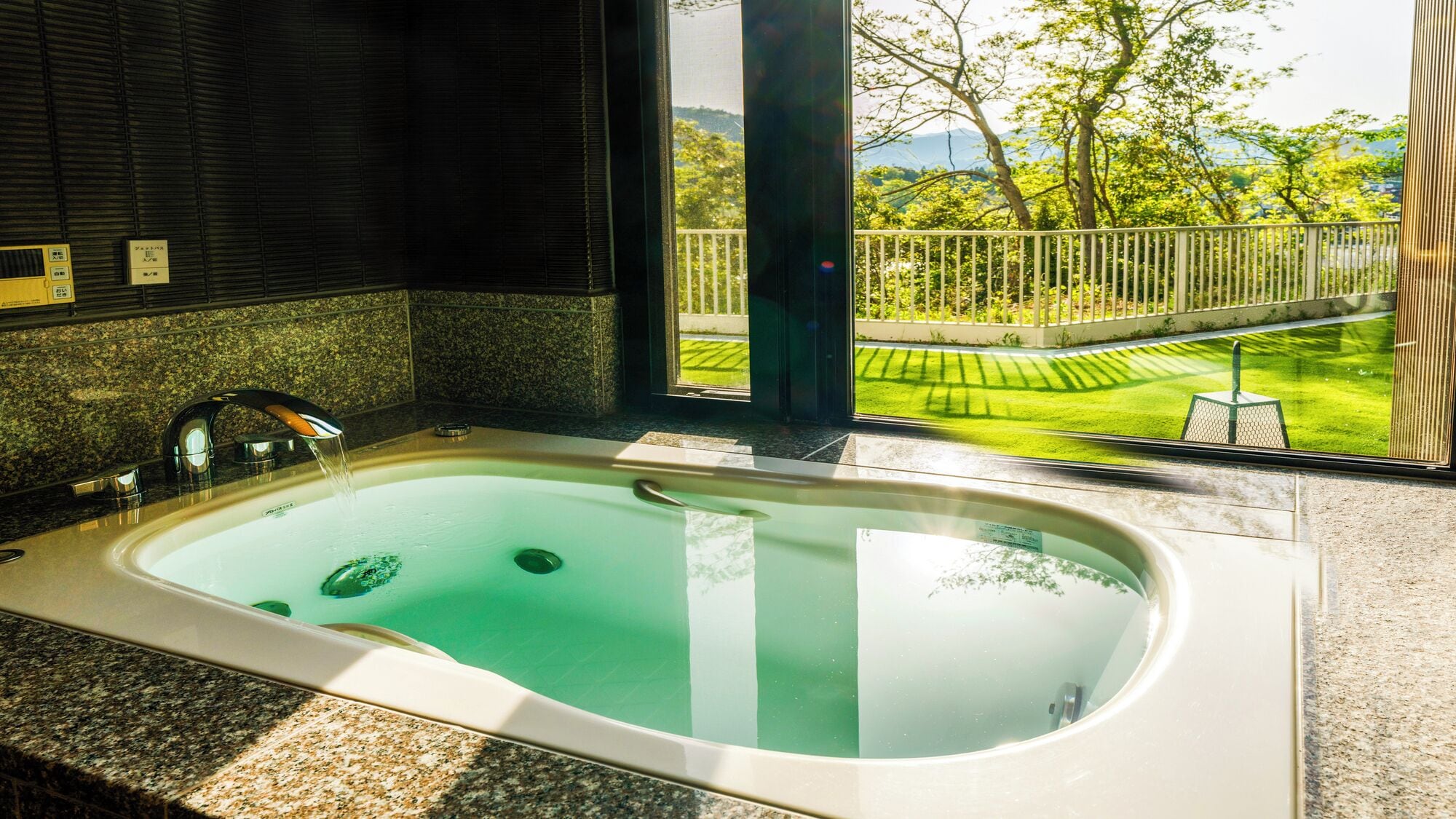 Un baño con una bañera amplia, encimera de granito y vistas a un césped y árboles bien cuidados.