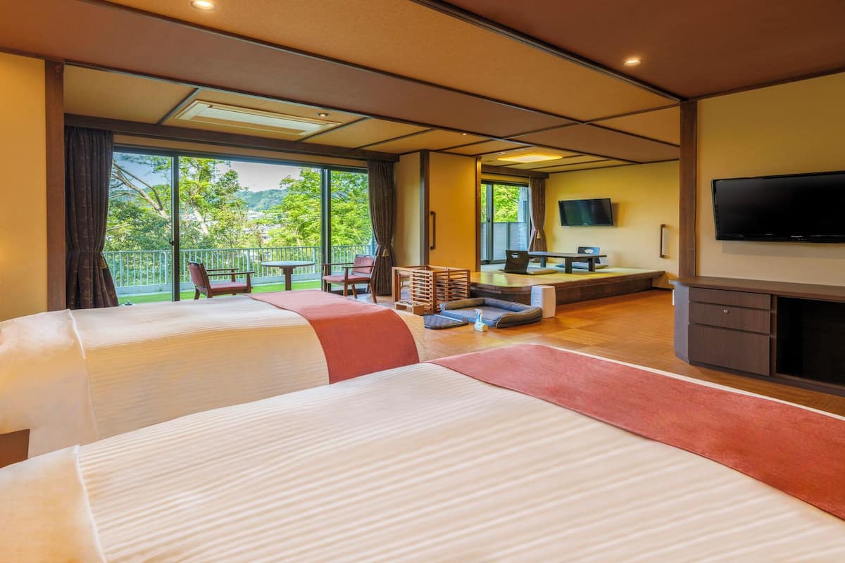A hotel room with two beds, a TV, a desk with a chair, and a balcony with a view of greenery.