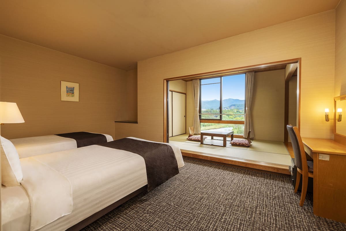 A hotel room with two beds, a desk, a chair, a window with a view of mountains, and a small table with a cushion.