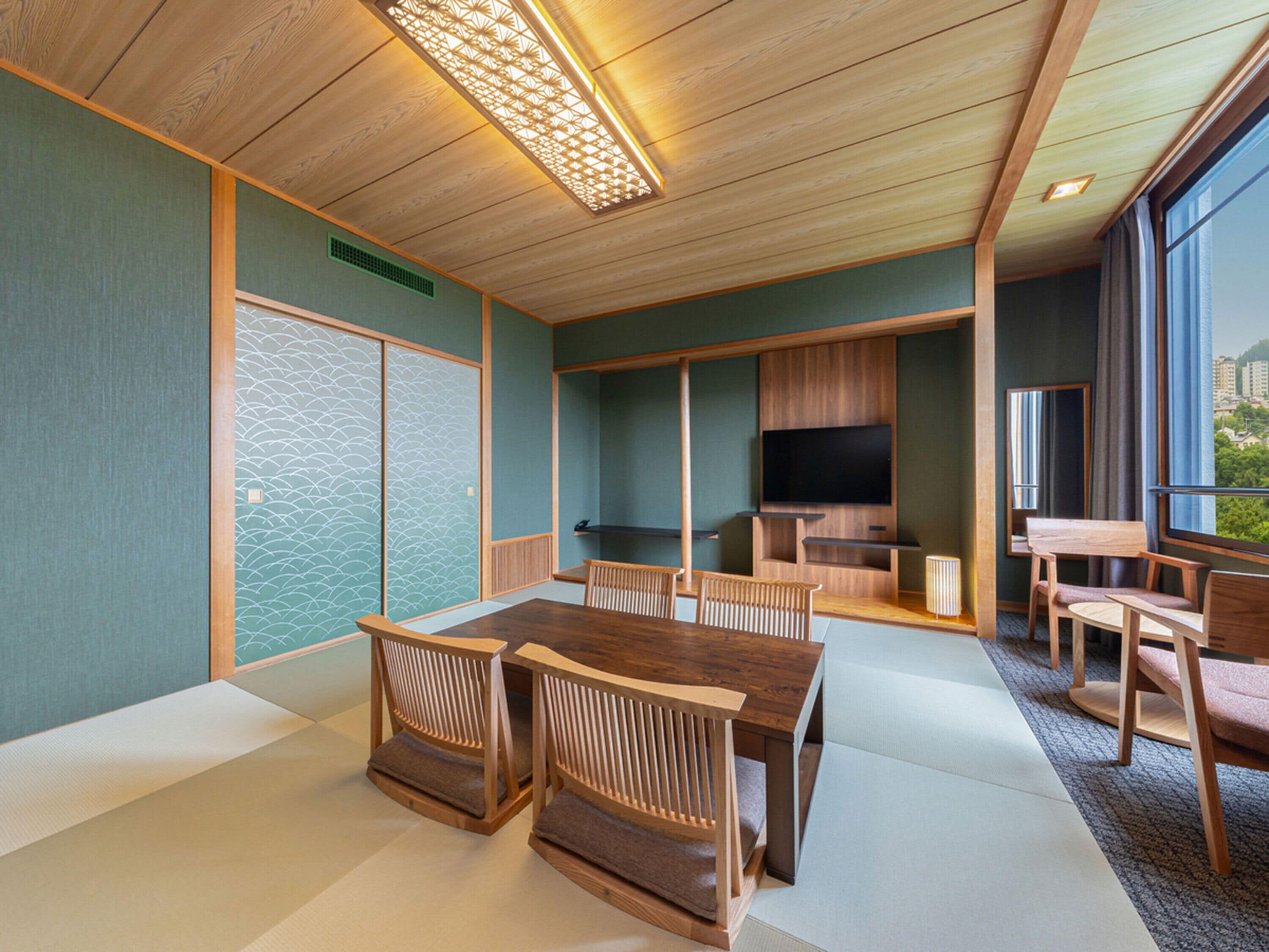 A modern interior with a wooden dining table, chairs, a flat-screen TV, and a large window offering a view of the city.