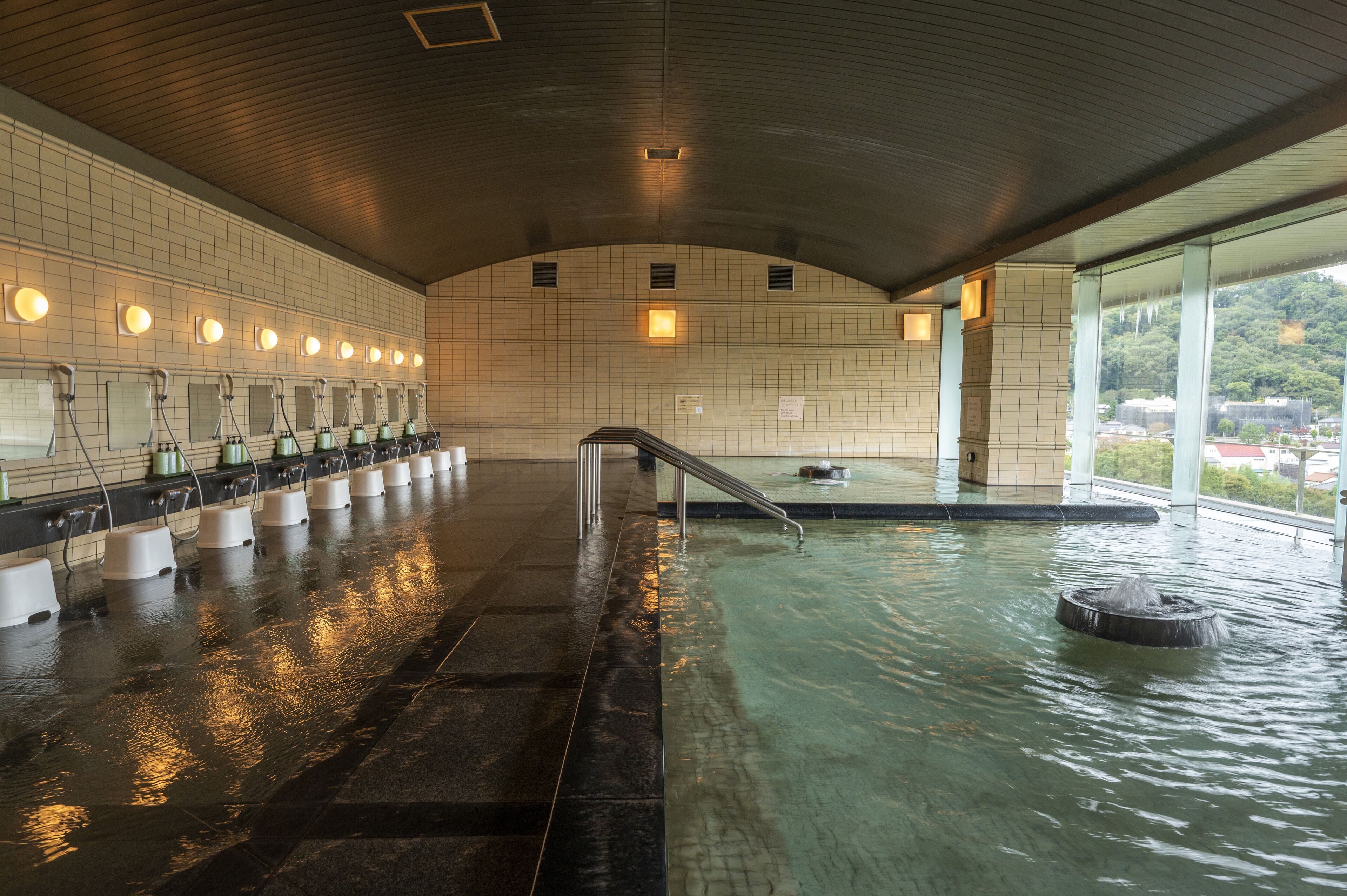 A public bath with multiple changing rooms, a central pool, and a view of a green hillside.