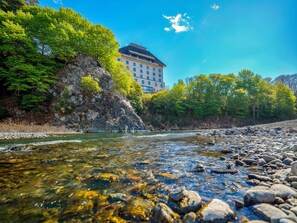 A river with clear water, surrounded by rocks and greenery, with a building in the background. - Kamenoi Hotel Ome (Ome)