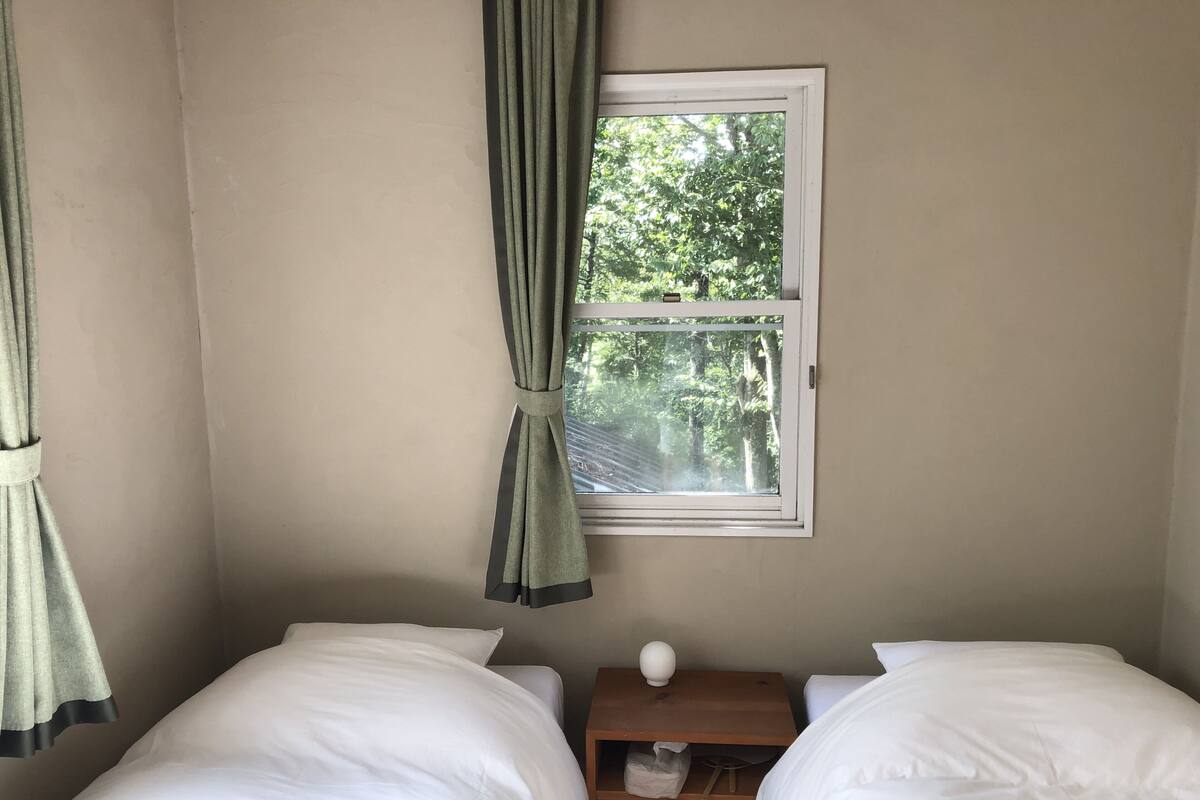A room with two single beds, a window with curtains, a small wooden nightstand, and a view of trees outside.