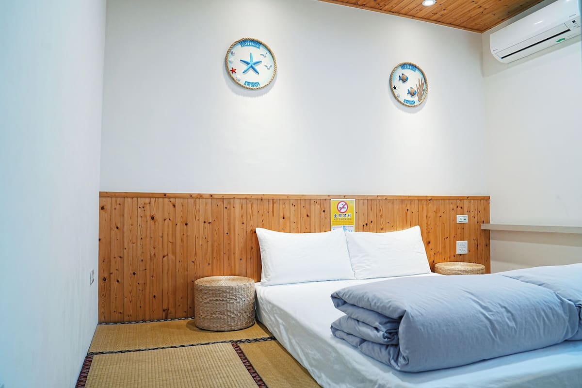 Une chambre d’hôtel avec une tête de lit en bois, deux horloges murales, un lit avec une literie bleue, un panier en osier et une petite table ronde.