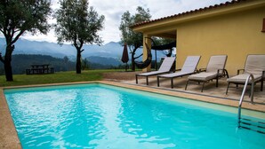 Una piscina con sillas de descanso y una mesa de picnic, rodeada de árboles y montañas al fondo.