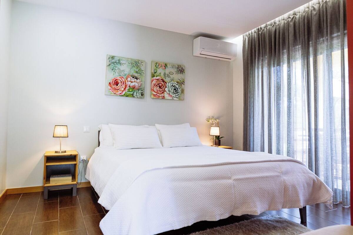 A hotel room with a large bed, two bedside tables with lamps, floral artwork on the wall, an air conditioning unit, and a view through sheer curtains.