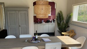 Un comedor con mesa de madera, sillas blancas, lámpara colgante de ratán y vista a la cocina a través de un arco.