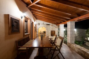 A covered outdoor dining area with a long wooden table and chairs, stone walls, and a stone fireplace.