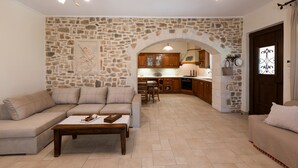 A living room with a stone wall, a sofa, a coffee table, and a kitchen in the background. - Private Villa with Pool & Jacuzzi | Organic Garden | Ideal for Families (RETHYMNO)