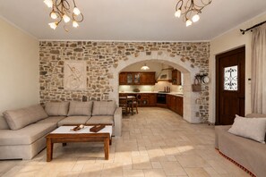 A living room with a stone wall, a sofa, a coffee table, and a kitchen in the background. - Private Villa with Pool & Jacuzzi | Organic Garden | Ideal for Families (RETHYMNO)